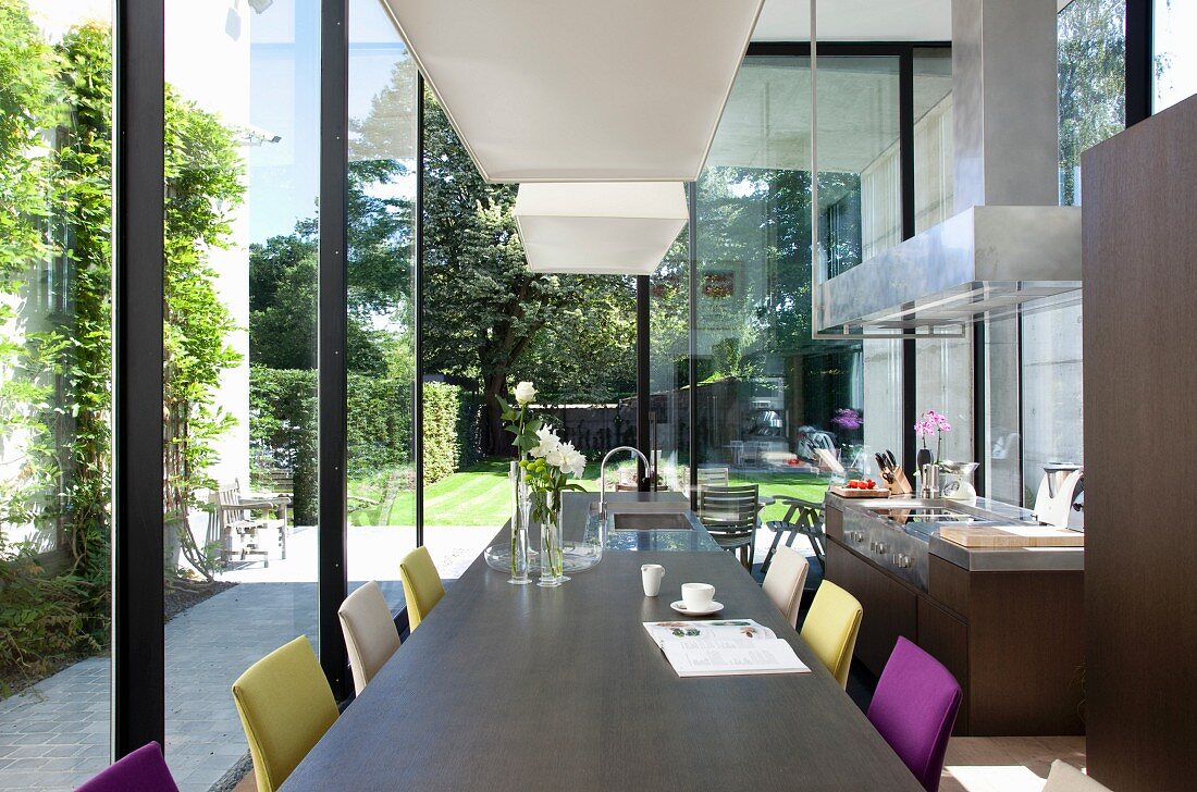 Lange Tafel und farbige Polsterstühle vor raumhohen Terrassenfenstern in offener Küche
