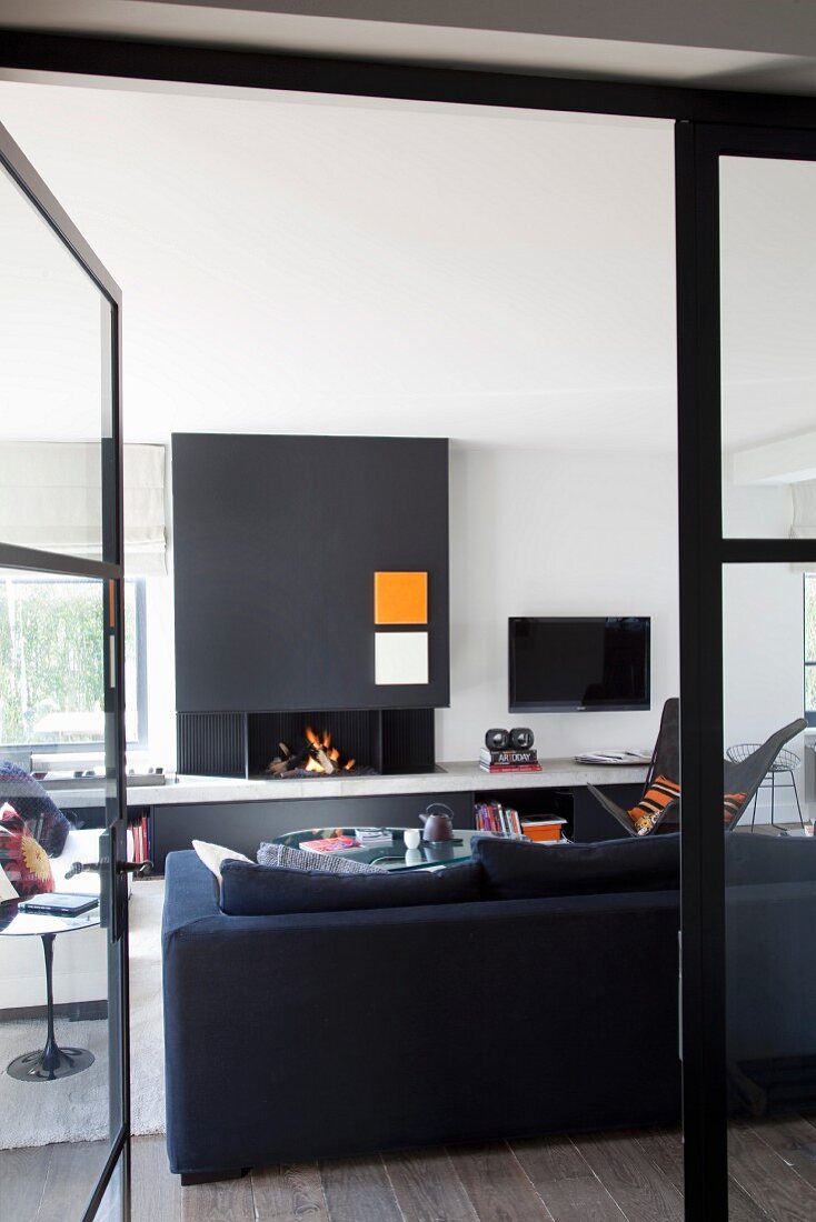 Blick durch offene Tür auf Rückseite eines schwarzen Sofas vor Kamin in modernem Wohnraum