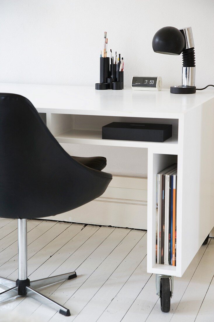 Weissglänzender Schreibtisch mit schmalen Fächern auf Rollen und schwarzer Lederdrehsessel auf weißem Dielenboden