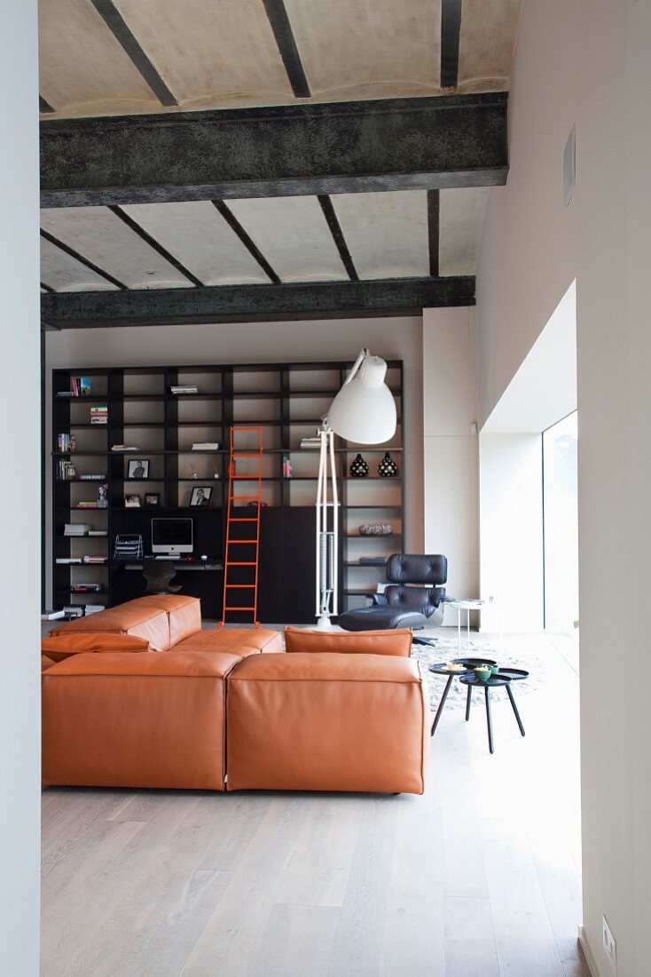 Loft apartment living room with old concrete ceiling on heavy steel joists, black, industrial-style shelving with bright orange ladder and leather couch in same shade of orange