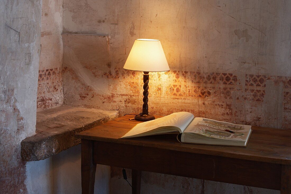 Tischlampe und aufgeschlagenes Buch auf Holztisch vor Wand mit verblasster Schablonenmalerei