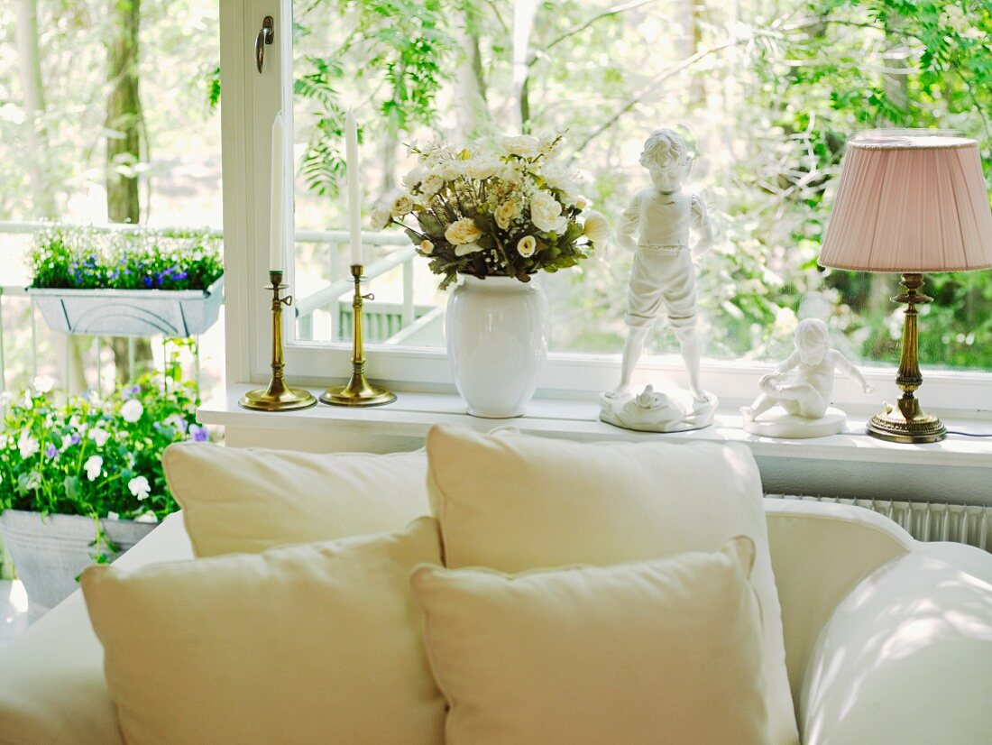 Kissen auf weißem Polstersessel vor Fensterfront mit Blumendeko und Wohnaccessoires im Vintagestil