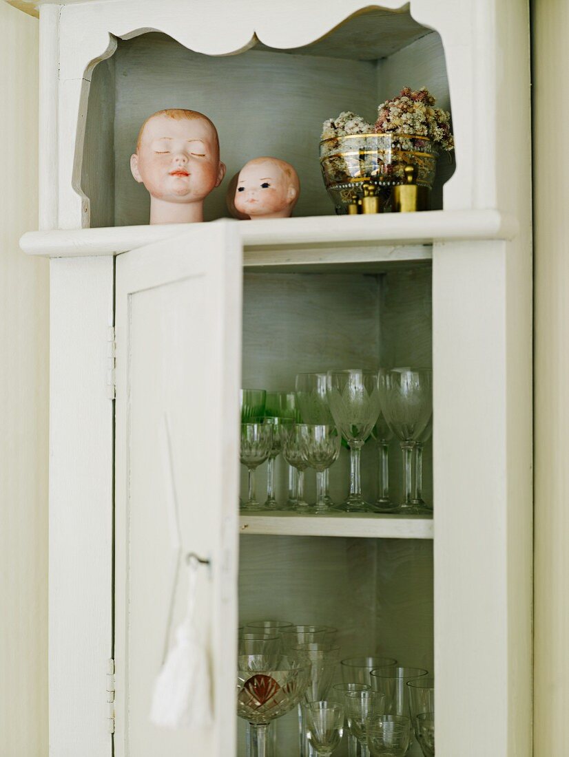 Glasses in romantic corner cupboard decorated with dolls' heads and floral potpourri