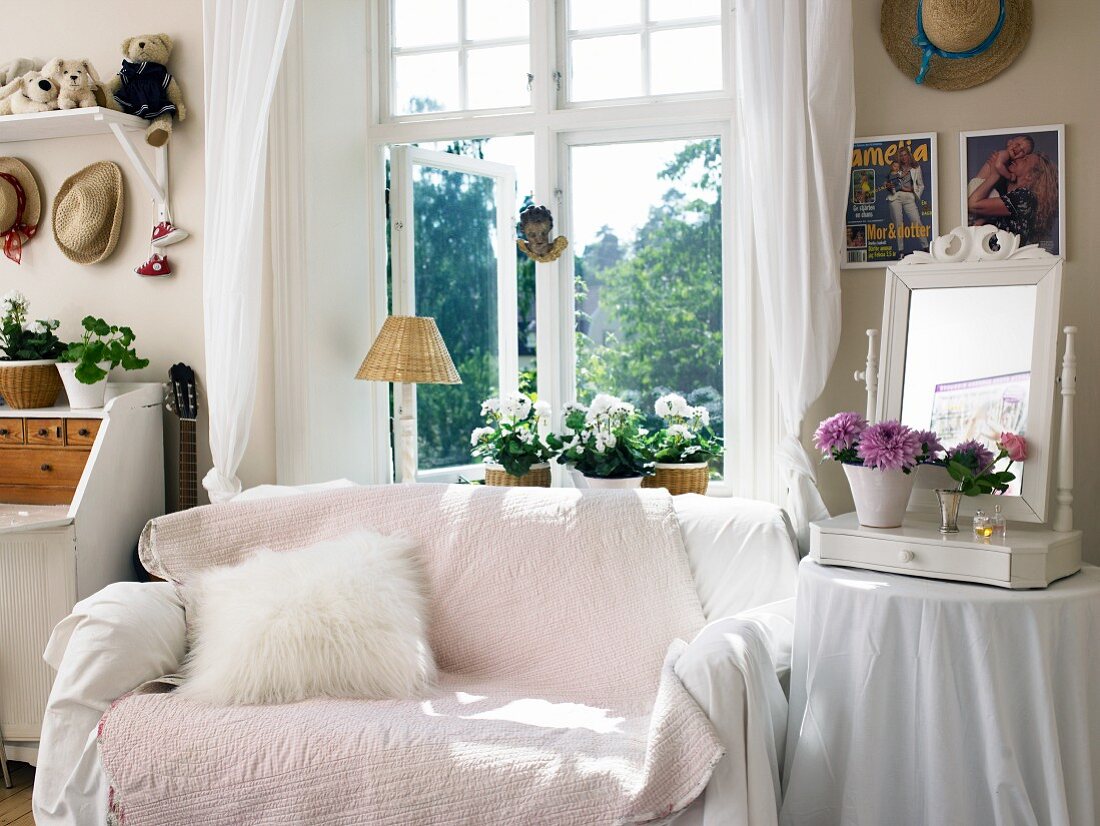 Sofa mit rosa Überwurf vor dem Fenster, daneben Beistelltisch mit Spiegel und Blumendeko
