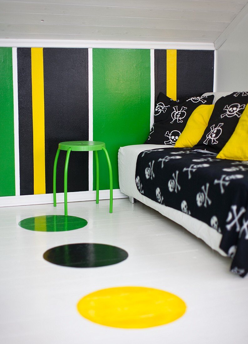 Sofa mit schwarzer Decke und Kissen und grüner Hocker in einem Dachzimmer mit bunt gestreifter Wand