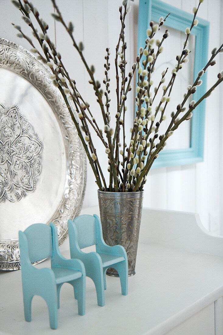 Metal vase with pussy willow branches next to miniature chairs on a white sideboard