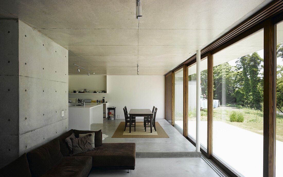 Exposed concrete in open-plan living space with kitchen and dining area in front of wide terrace doors with view of nature