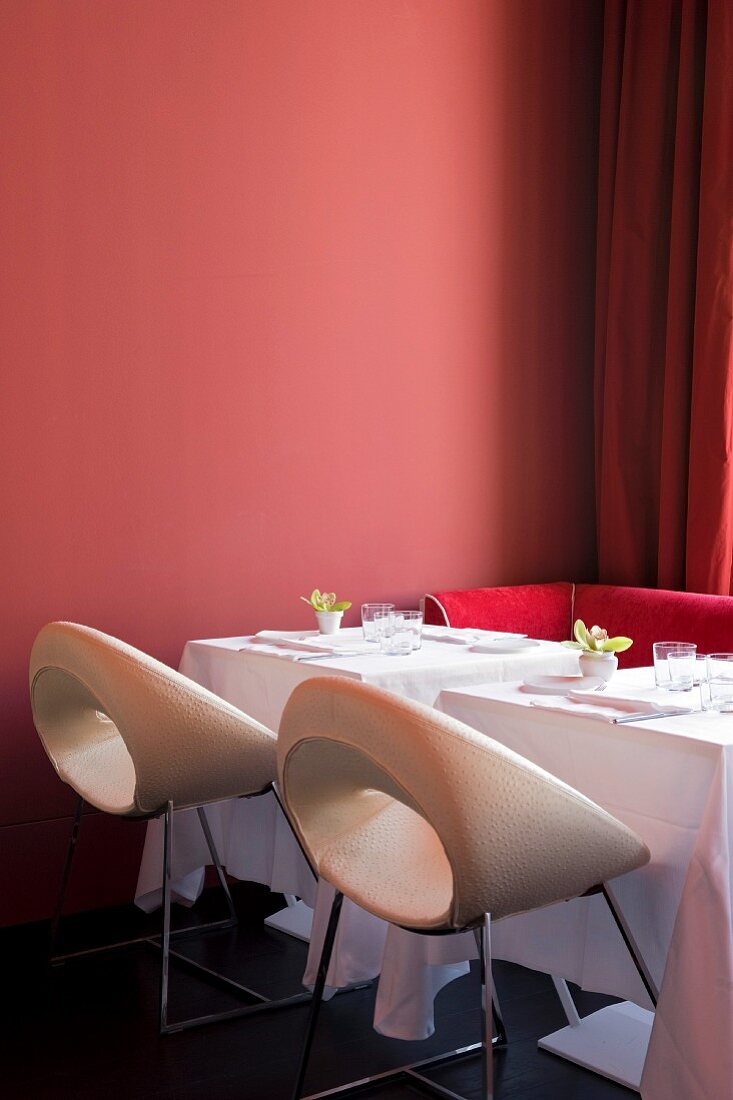 Gedeckte Zweiertische mit weissen und pinkroten Designerstühlen vor ziegelrot getönter Wand
