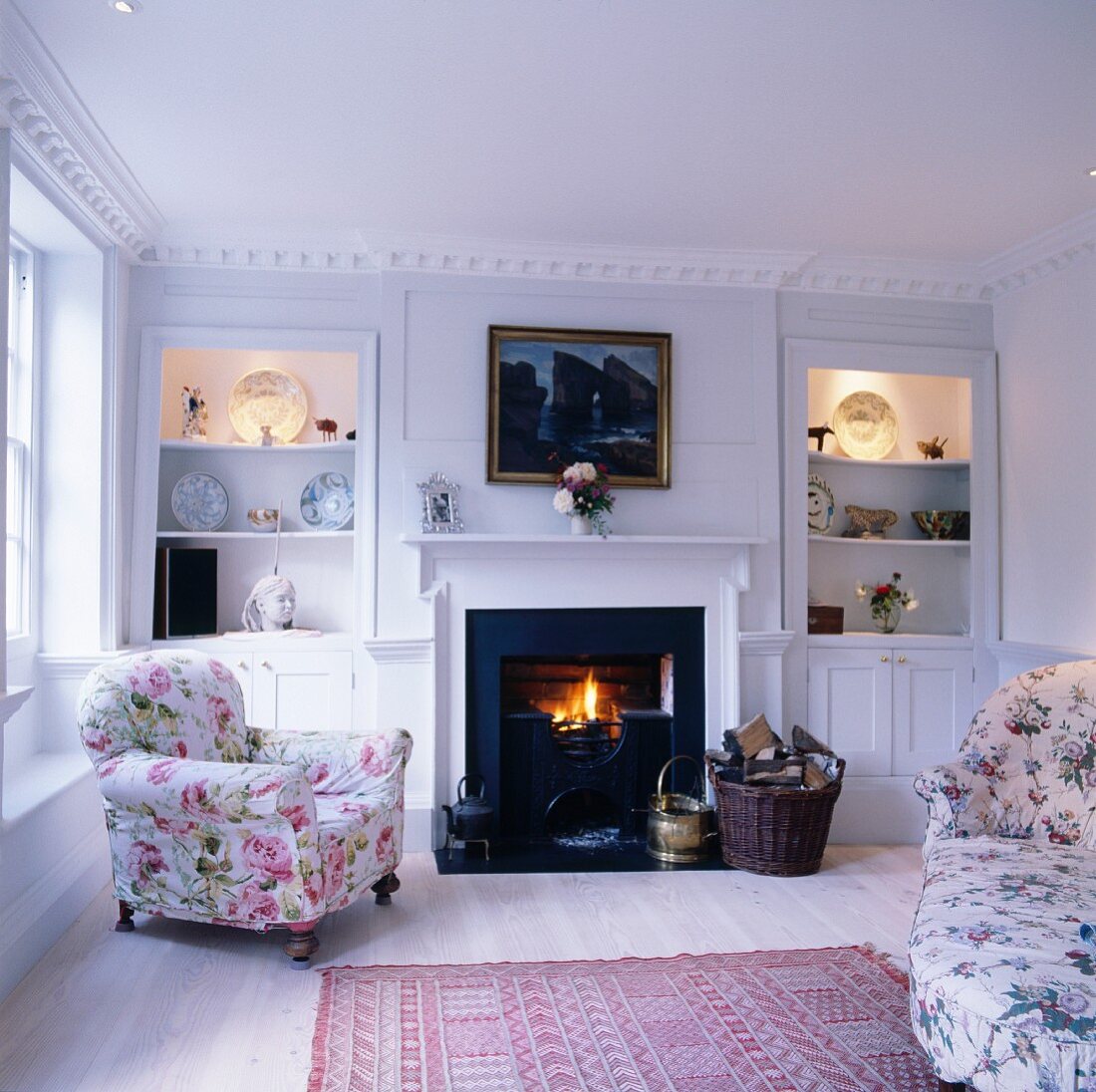 Wohnzimmer im romantischen Landhausstil mit floral gemusterten Polstermöbeln und Kaminfeuer zwischen beleuchteten Einbauregalen
