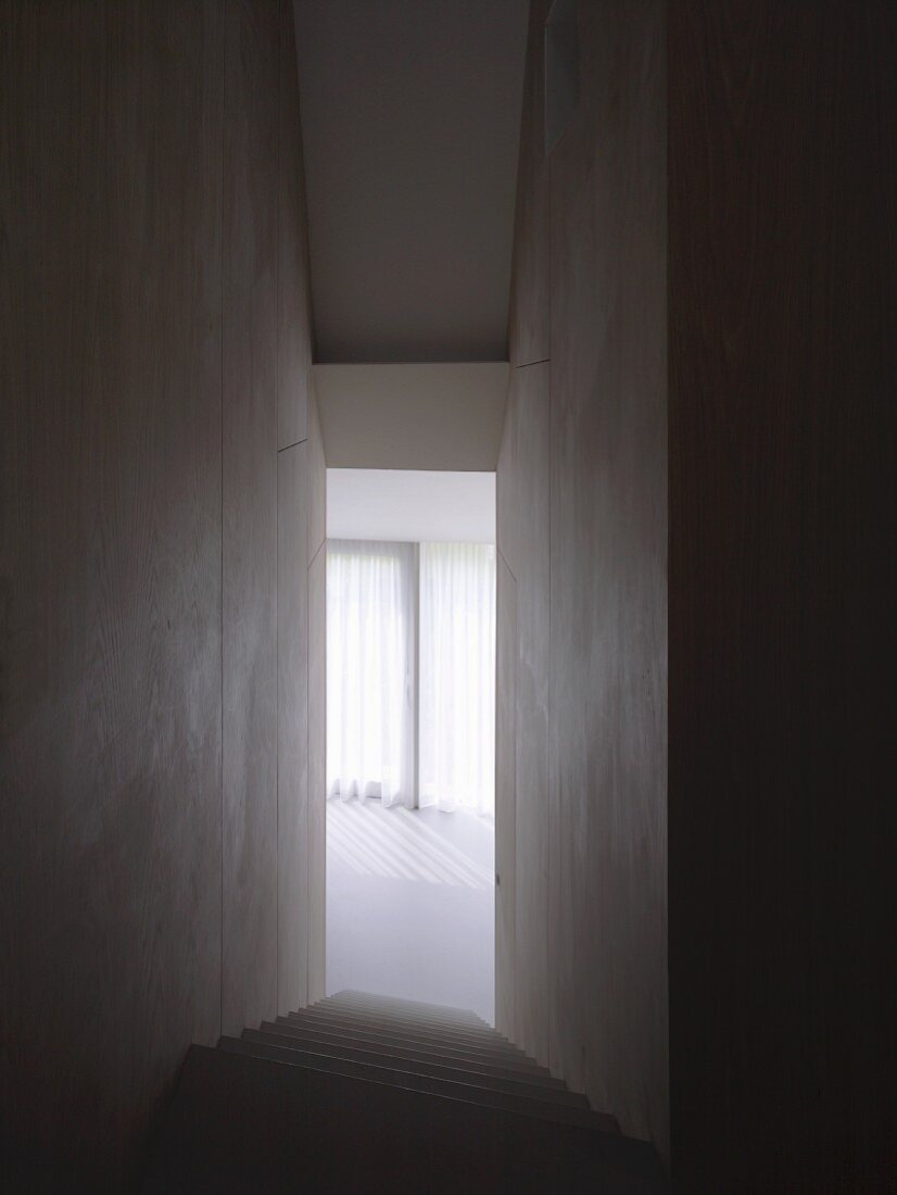 Schmaler, hoher Treppenabgang ohne Handlauf und Blick in sonnigen Raum mit Vorhang aus Vertikallamellen