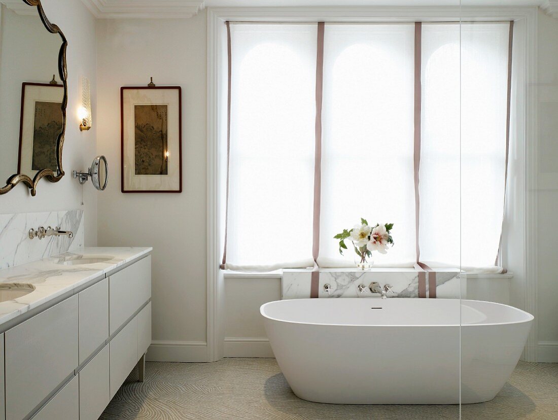 Freistehende Badewanne vor Fenster mit Rollovorhang in elegantem Bad mit Marmorplatten und Spiegel mit Zierrahmen