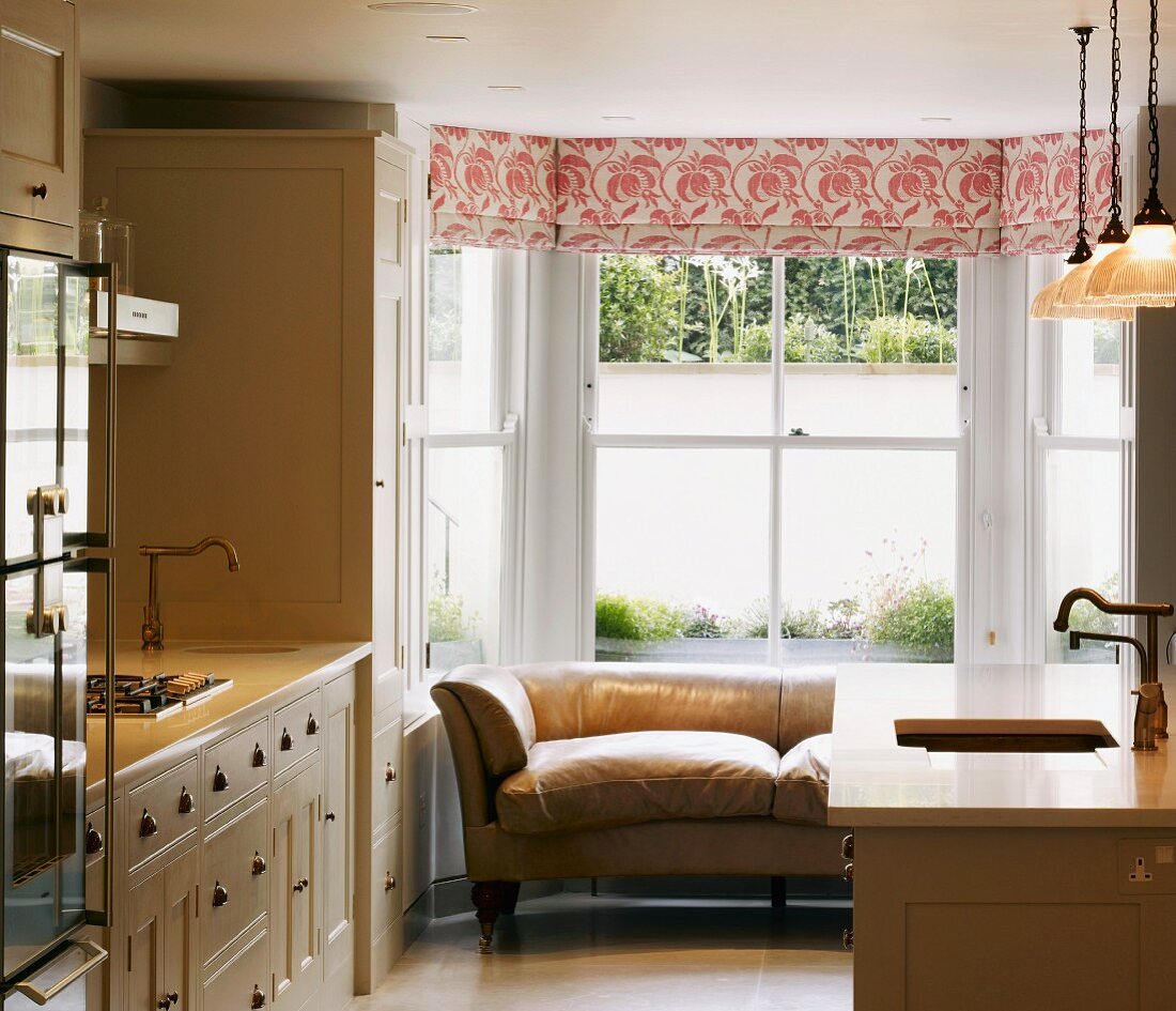 Cream-coloured, retro-style country house kitchen with leather couch in bay window and floral Roman blind