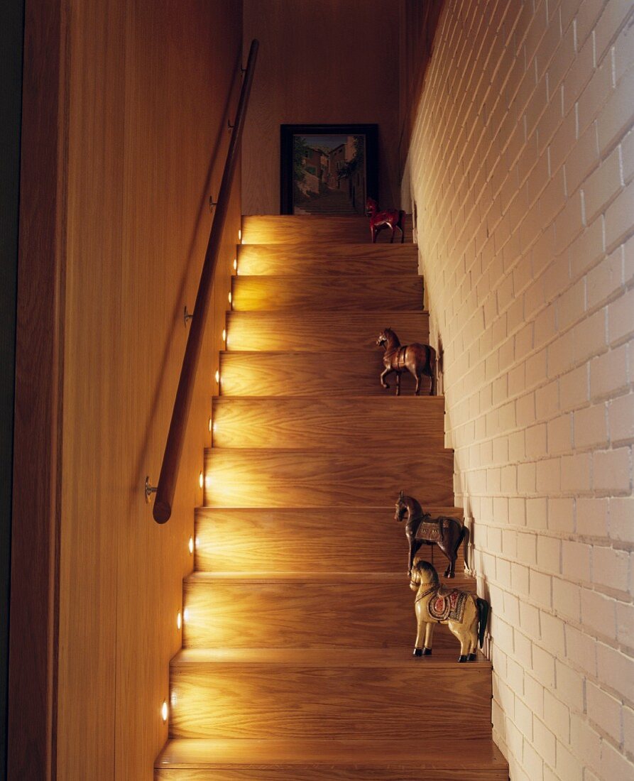 Sammlung von bemalten Holzpferden auf den beleuchteten Stufen eines Treppenaufgangs mit weiss gestrichener Backsteinwand