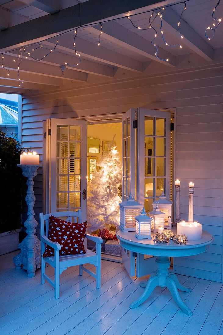 Festliche Stimmung auf Veranda mit Laternen und Kerzenständern auf Tisch vor offener Wohnzimmertür und Blick auf beleuchtetem Weihnachtsbaum