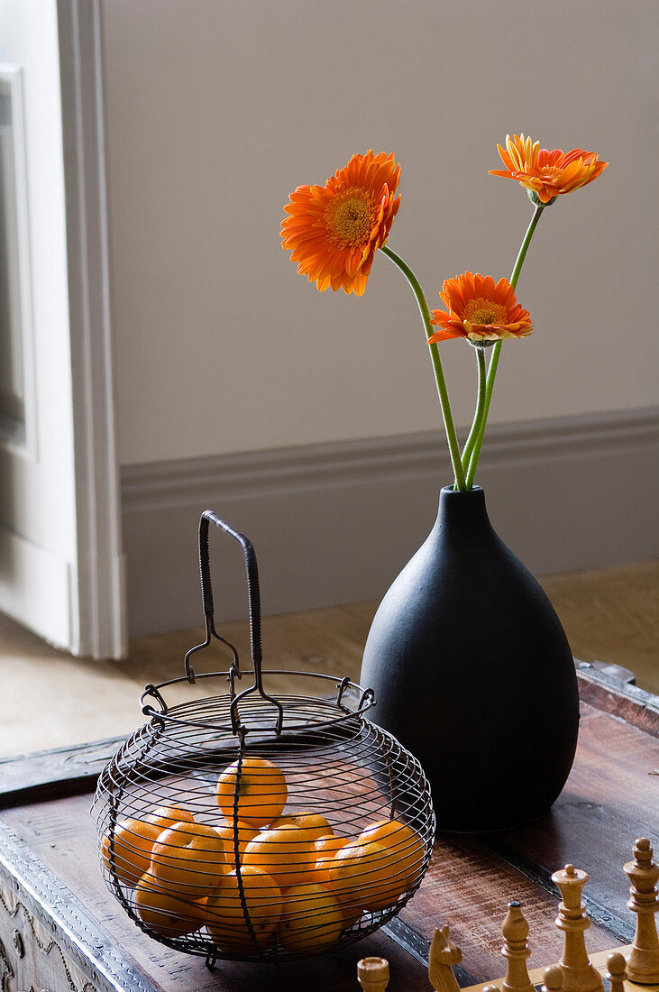 Schwarze Vase mit orangefarbenen Gerbera und antiker Drahtkorb mit Obst auf antikem Holztisch