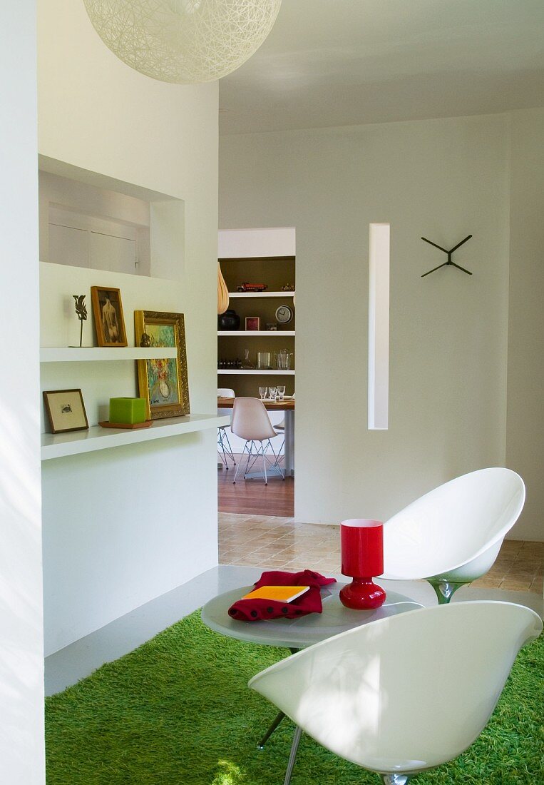 Zwei Schalenstühle aus weißem Kunststoff auf grünem flokatiartigem Teppich in offenem Wohnraum mit Blick auf Esstisch