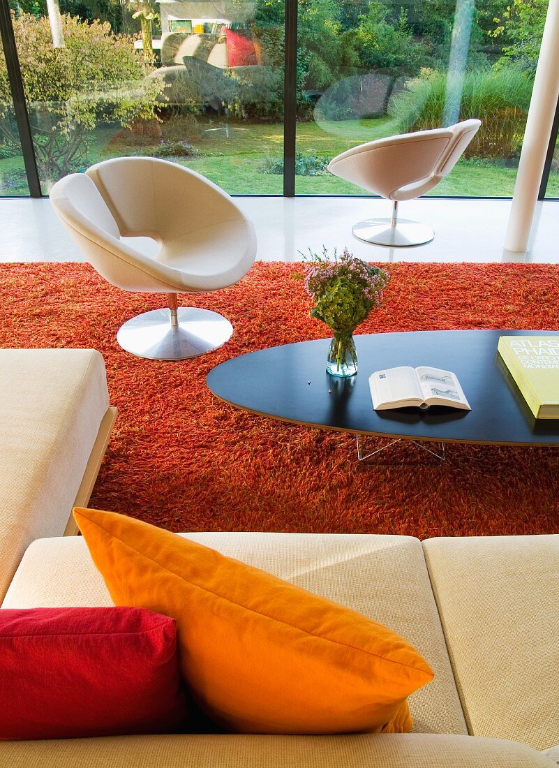 Classic coffee table and white, designer swivel chair on red, flokati-style rug in contemporary building with glass facade and view of garden