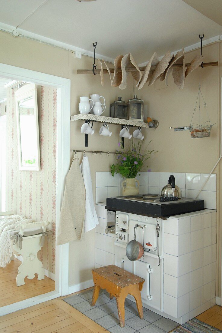 Holzschemel vor rustikalem Küchenofen mit weissen Fliesen in Zimmerecke neben Durchgang und Blick auf Sitzbank aus weißem Holz