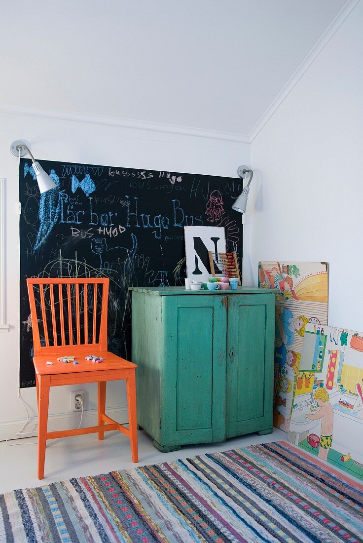 Orangefarbener Holzstuhl und grünes Schränkchen vor Kreidetafel an der Wand und Flickenteppich