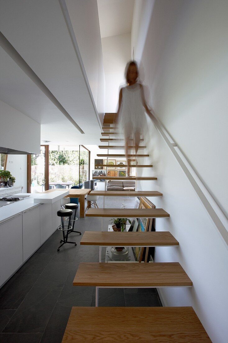 Woman walking down minimalist staircase into open-plan living area with slate floor and large terrace windows