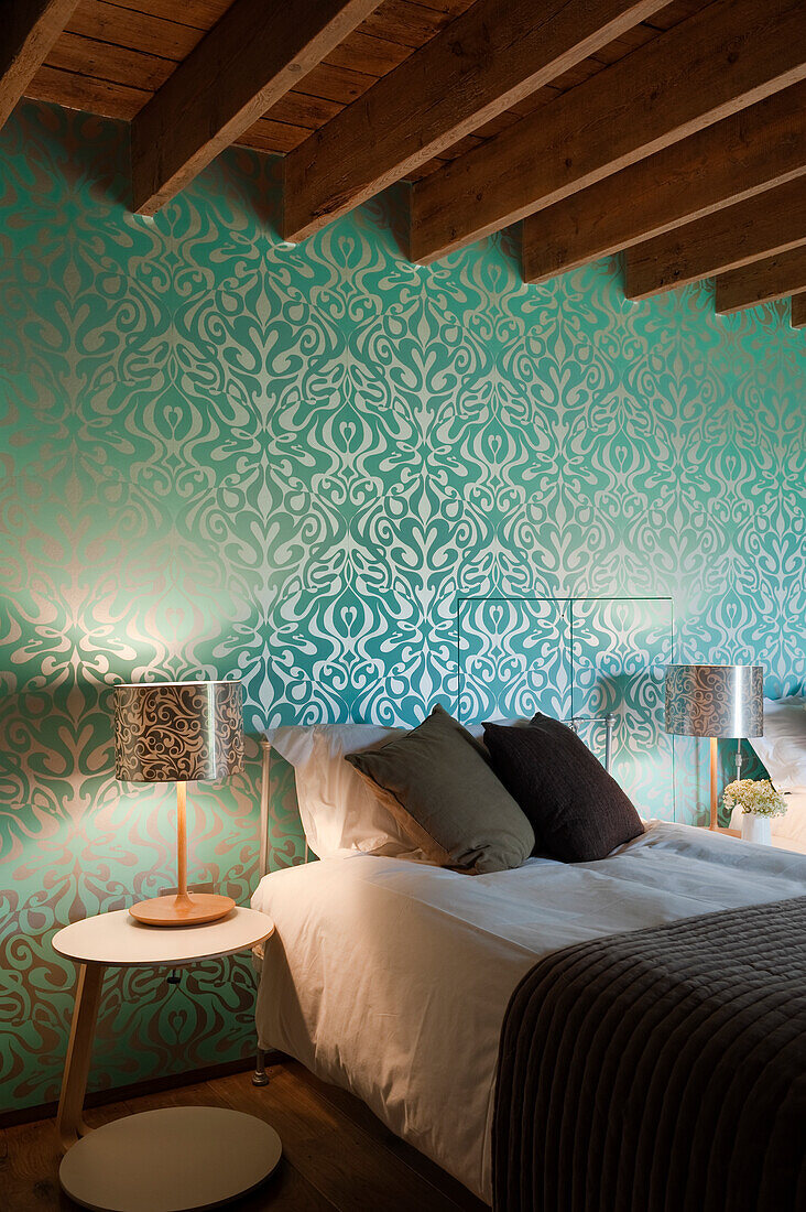 Patterned wallpaper and wooden beams in the bedroom with bedside lamp