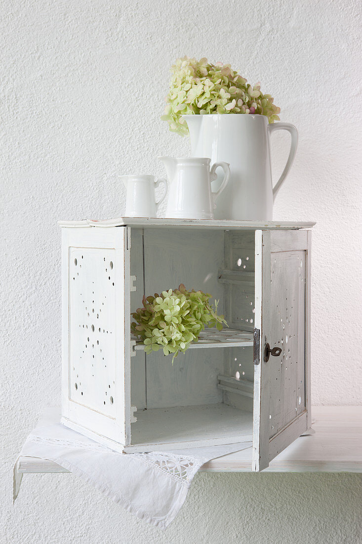 Small white cabinet with open door, set of jugs and flowers