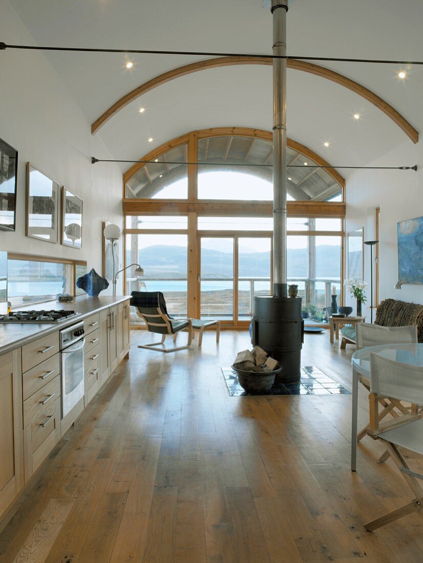Long kitchen unit and central wood-burning stove in open living space with arched ceiling and view of coastal landscape