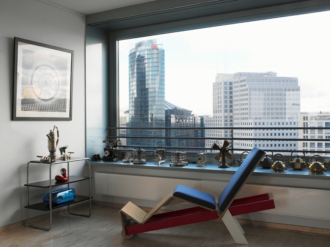 Gepolsterter Holzliegestuhl und Regal im Retrostil vor dekoriertem Panoramafenster mit Blick auf Berliner Hochhaus-Kulisse