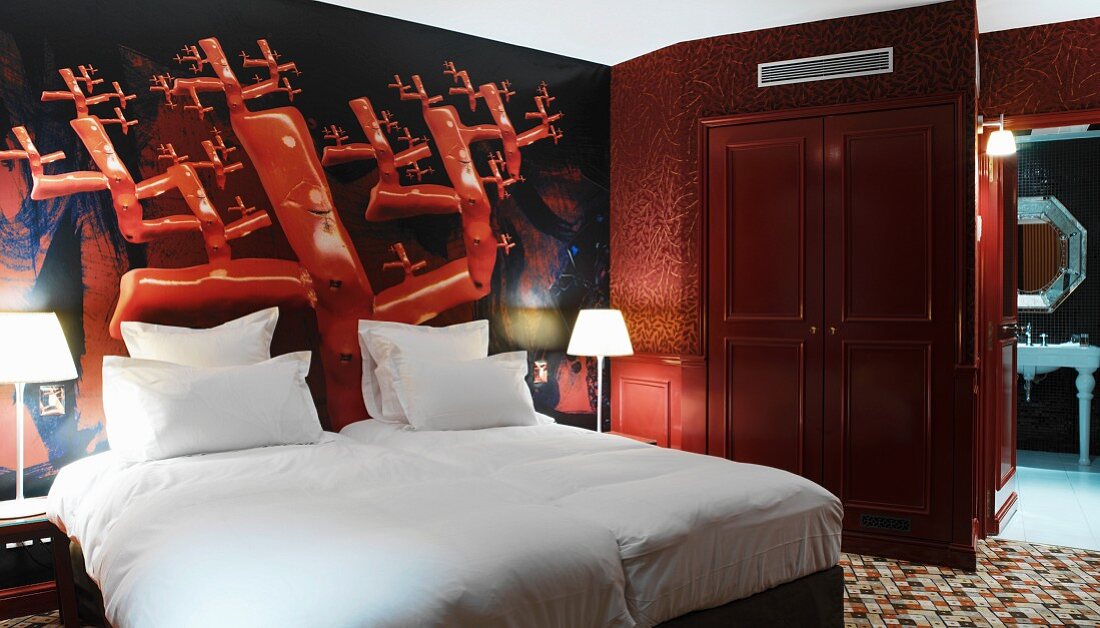 Plakative Wandbemalung in Knallrot hinter blütenweissem Bett in Hotelzimmer mit Bad ensuite
