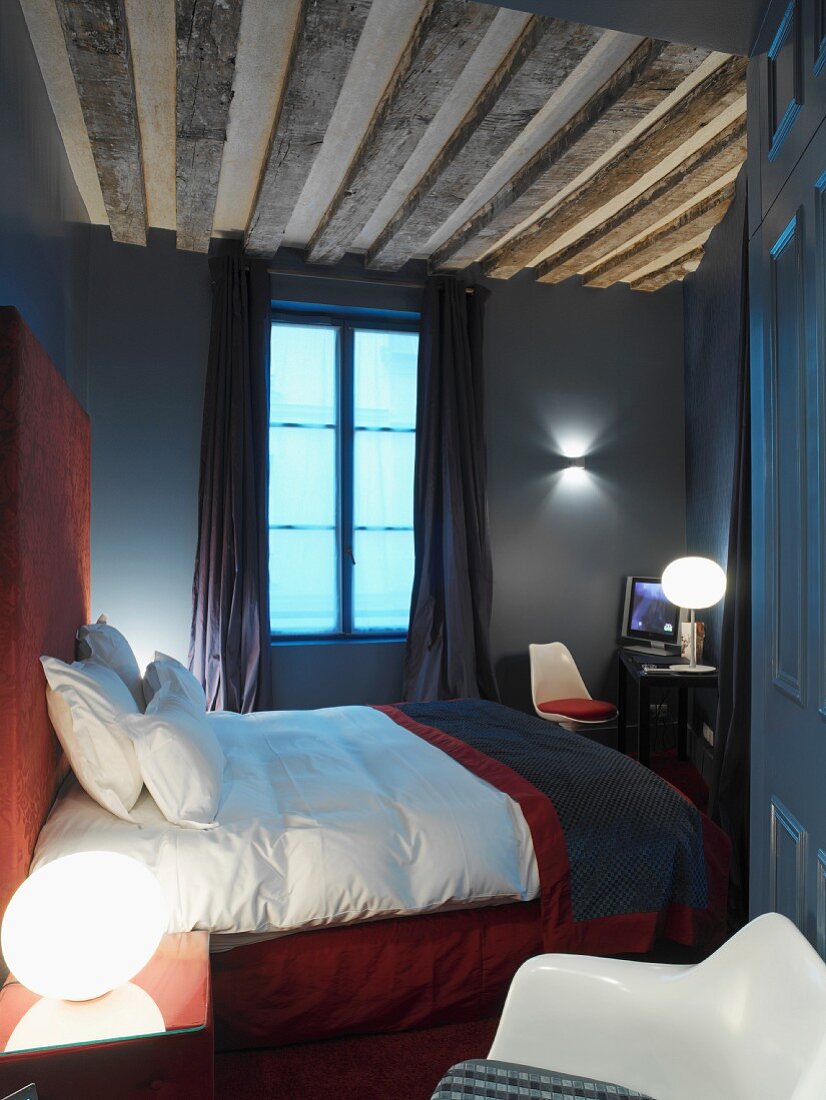 Doppelbett mit rotem Kopfteil in französischem Hotelzimmer in dunklen Farben und offener Holzbalkendecke