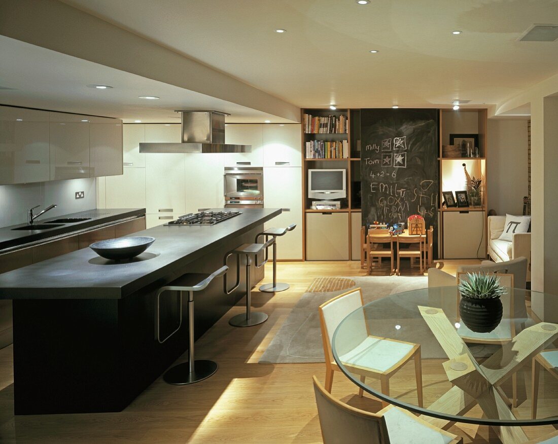 Moderner, offener Wohnraum mit langer Küchenbar, Essplatz mit Glastisch und Kindertisch vor raumhoher Kreidetafel