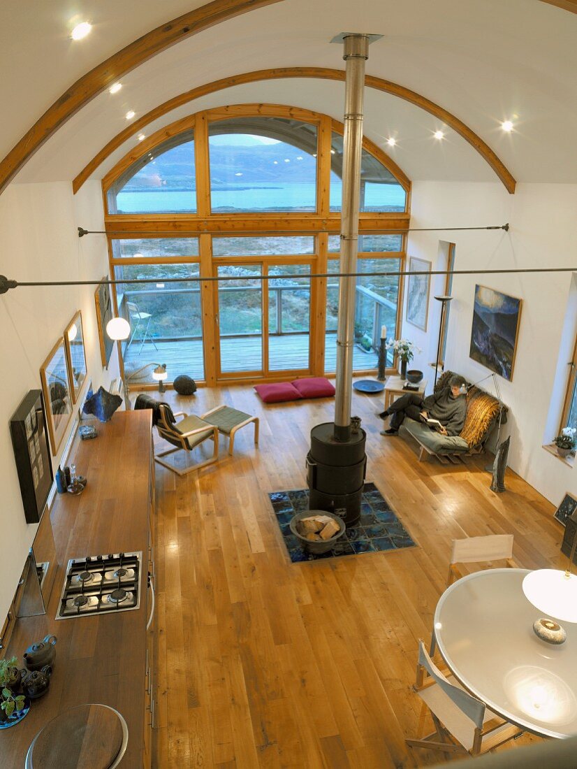 Offener Wohnraum von oben mit zentralem Kaminofen unter hohem Tonnendach und Fensterfront mit Blick auf Holzterrasse