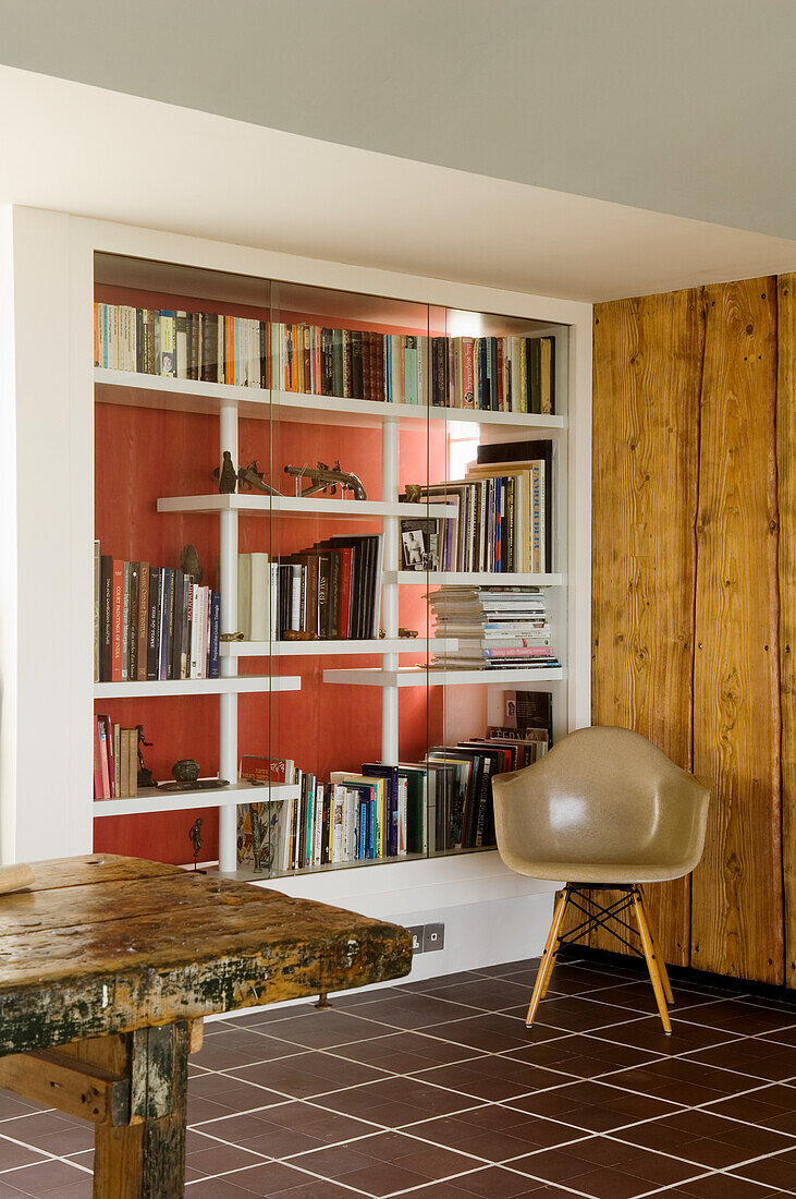 Open wall of books with red background, designer chair, rustic wooden table and tiled floor