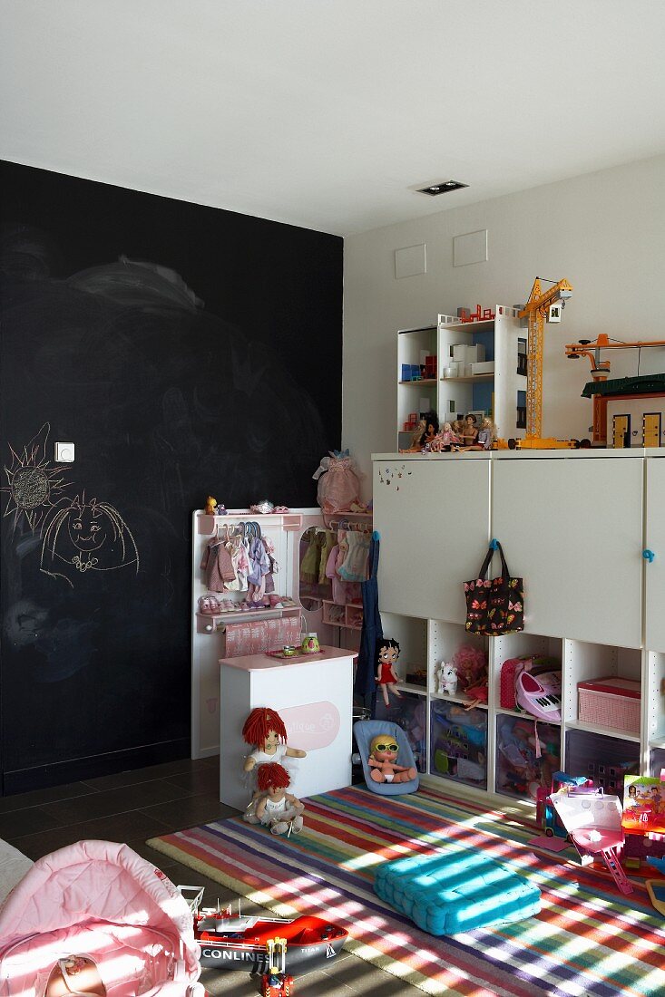 Schwarze Wand mit Kinderzeichnungen; davor weiße Kinderzimmermöbel, ein bunter Teppich und Mädchen-und Jungenspielzeug