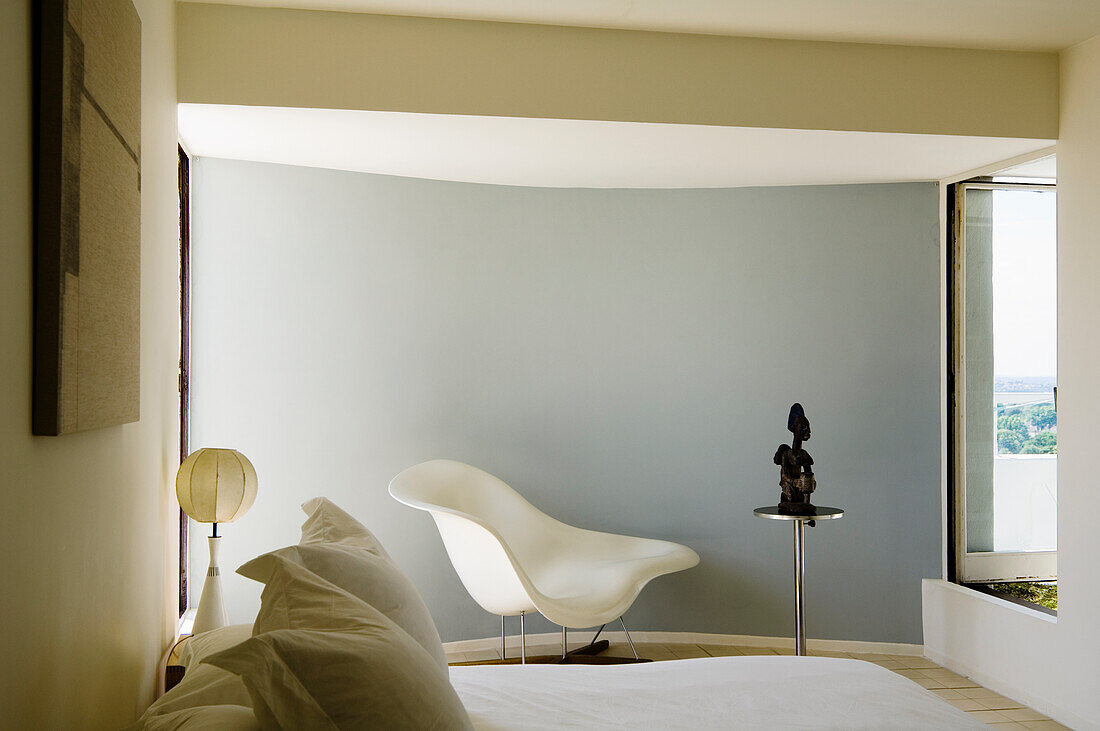 Minimalist bedroom with curved white lounge chair next to small side table and sculpture