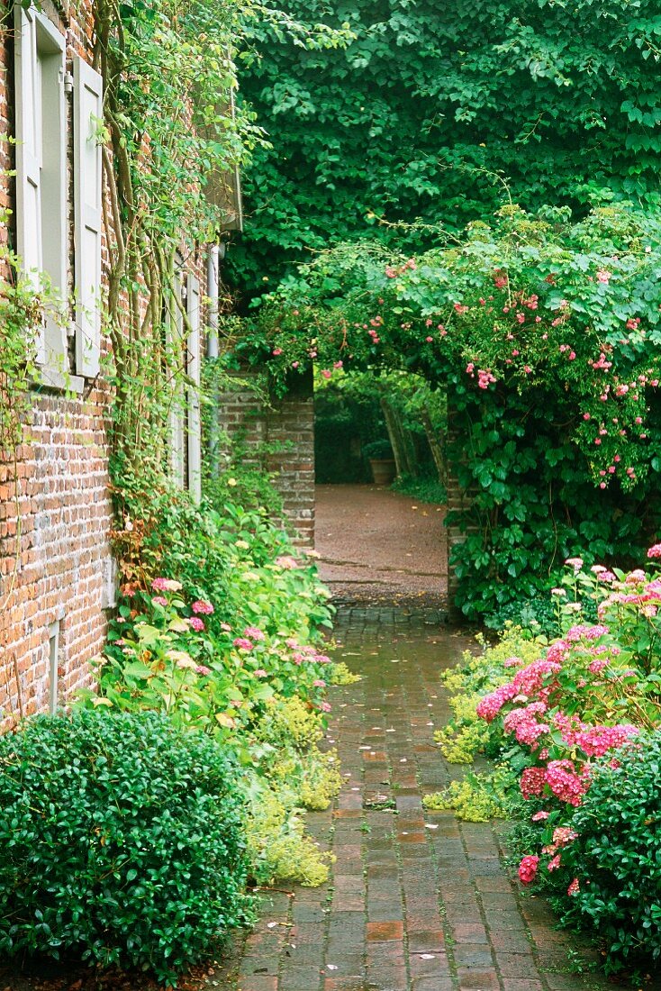 Rustic, paved garden path and roses next to house with climber-covered brick facade. Gateway leading to outdoor space beyond.