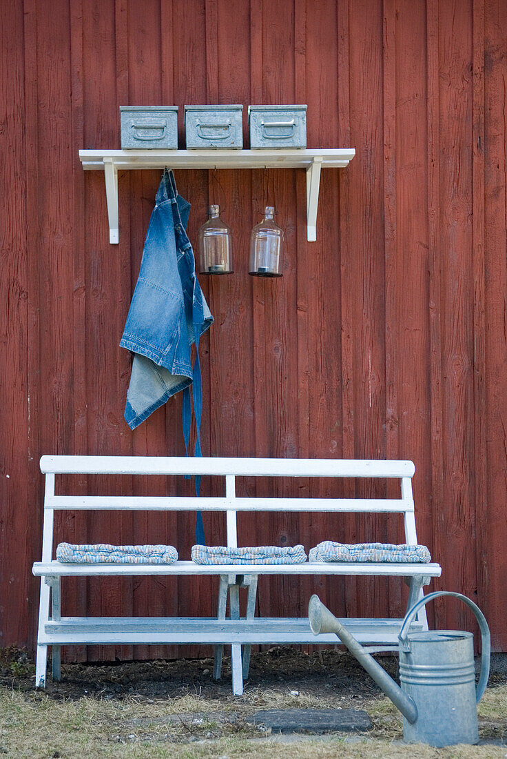 White garden bench with cushions, shelf, watering can and red wooden wall