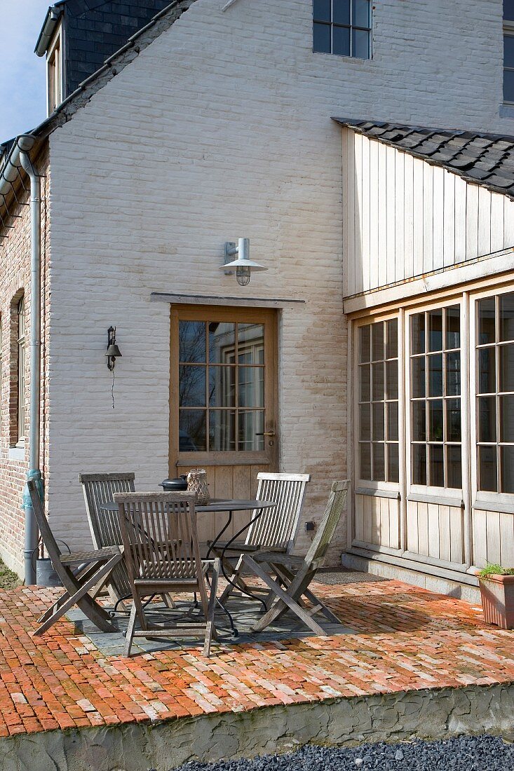 Seating area with folding wooden chairs on brick terrace in front of whitewashed brick house