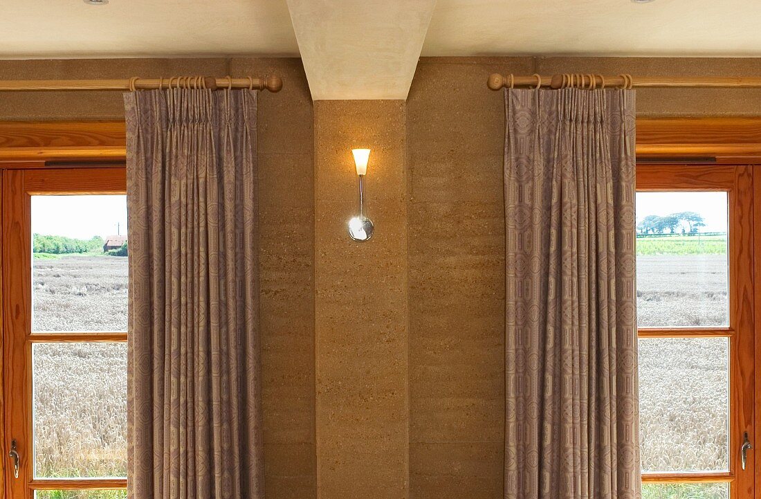 Kleine moderne Wandlampe zwischen Holzfenstern mit Vorhängen an Holzstangen