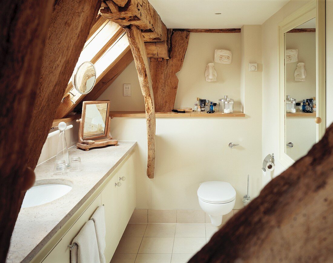 Niedriges, cremefarbenes Bad mit Marmorwaschtisch unter sichtbaren alten Dachbalken und Schrägverglasung