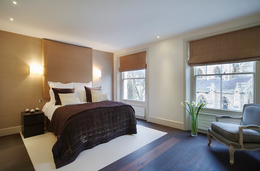 Schlafzimmer mit Faltjalousien vor grossen Fenstern, modernem Doppelbett und antikem Sessel