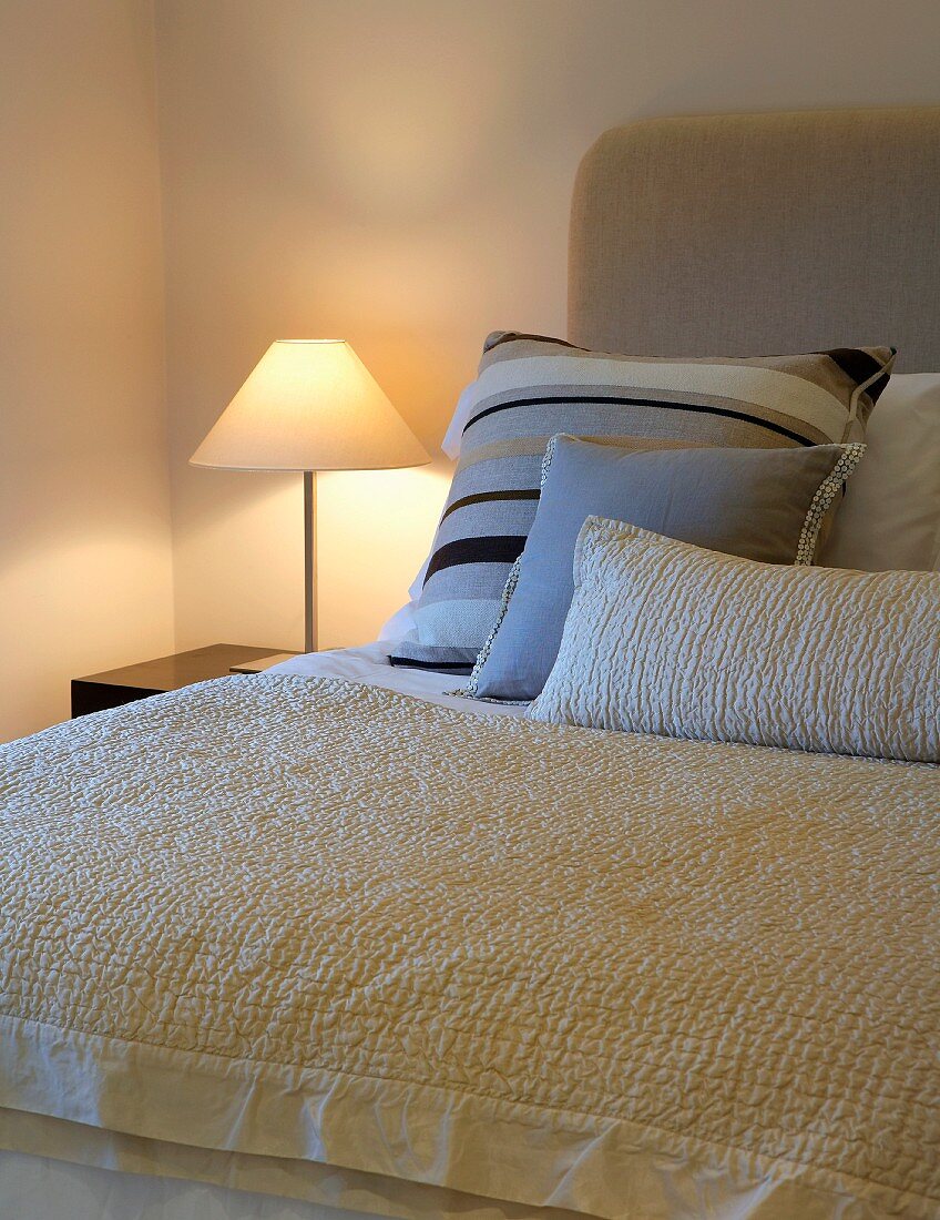 Scatter cushions and bedspread on double bed next to lit bedside lamp