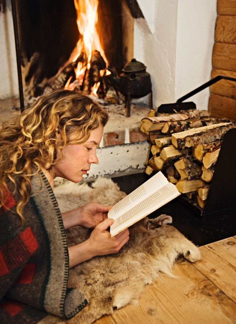 Frau vor offenem Kamin liegend und ein Buch lesend