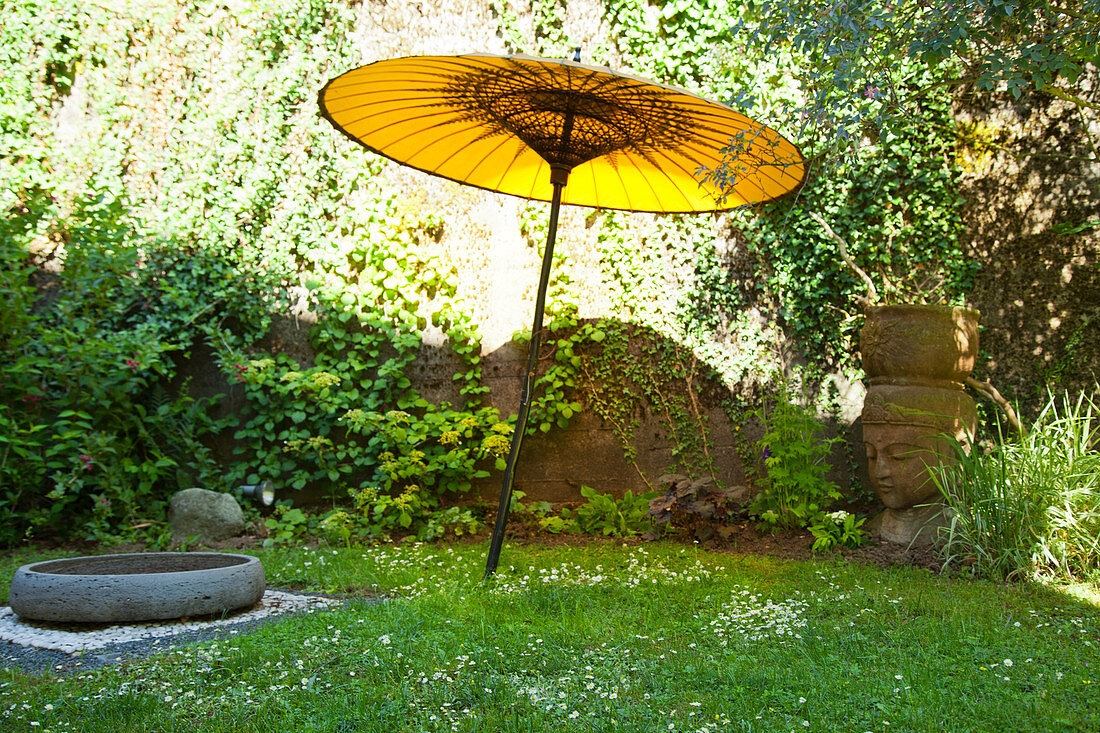 Gelber Sonnenschirm aus Papier, große Schale aus Stein und Buddhakopf auf einer Gartenwiese