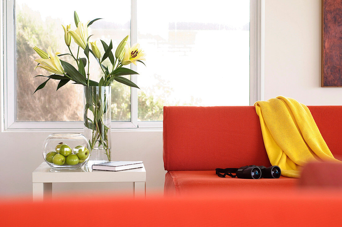 Lilienstrauss neben einem orangefarbenen Designer-Sofa mit gelber Decke und Fernglas