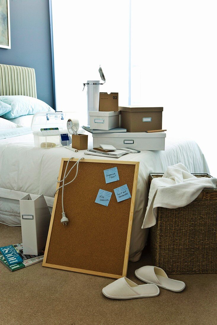 Double bed with storage boxes on top, a bulletin board leaning against it and a laundry basket; in front, white house slippers
