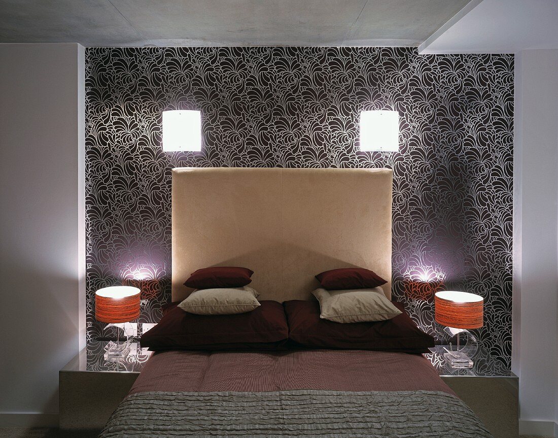 Stacked pillows on bed with high headboard and lamps on wall with patterned wallpaper