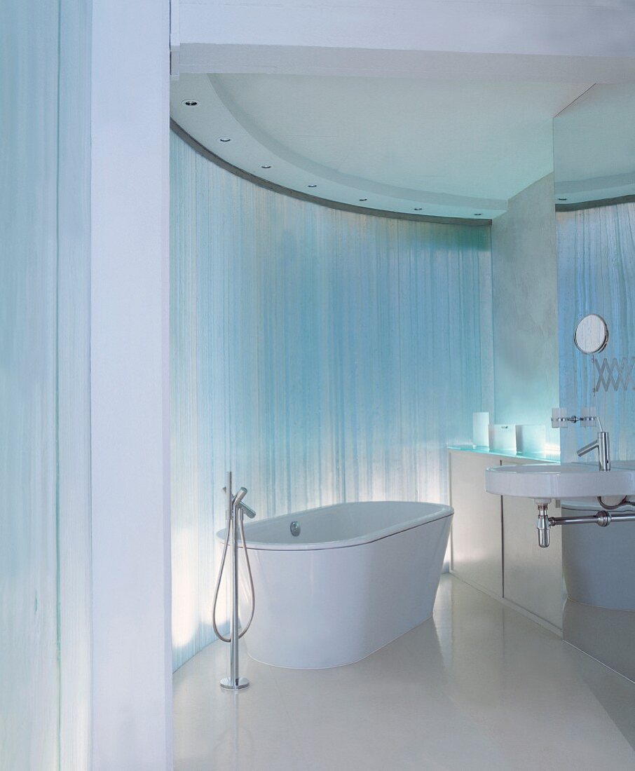 Free-standing bathtub with floor-mounted taps in front of curved, reflective wall