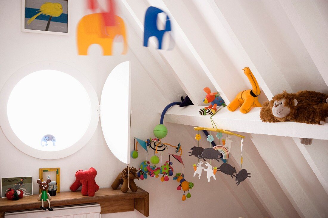 Bullaugenfenster, Kuscheltiere und bunte Mobile unter weiss gestrichener Dachkonstruktion im modernen Kinderzimmer