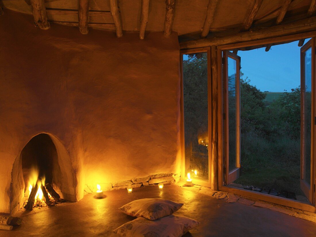 Brennender Kamin, Kerzenlicht und Bodenkissen vor offener Terrassentür in einfachem Lehmbau